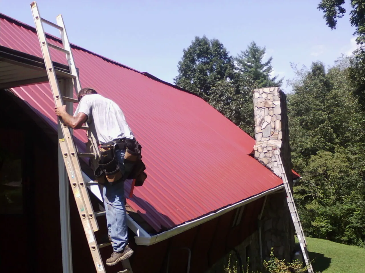 Roofer on a ladder beside a red metal roof and stone chimney — Soffit & Fascia Repair by Roofing Brothers in Saint Anthony